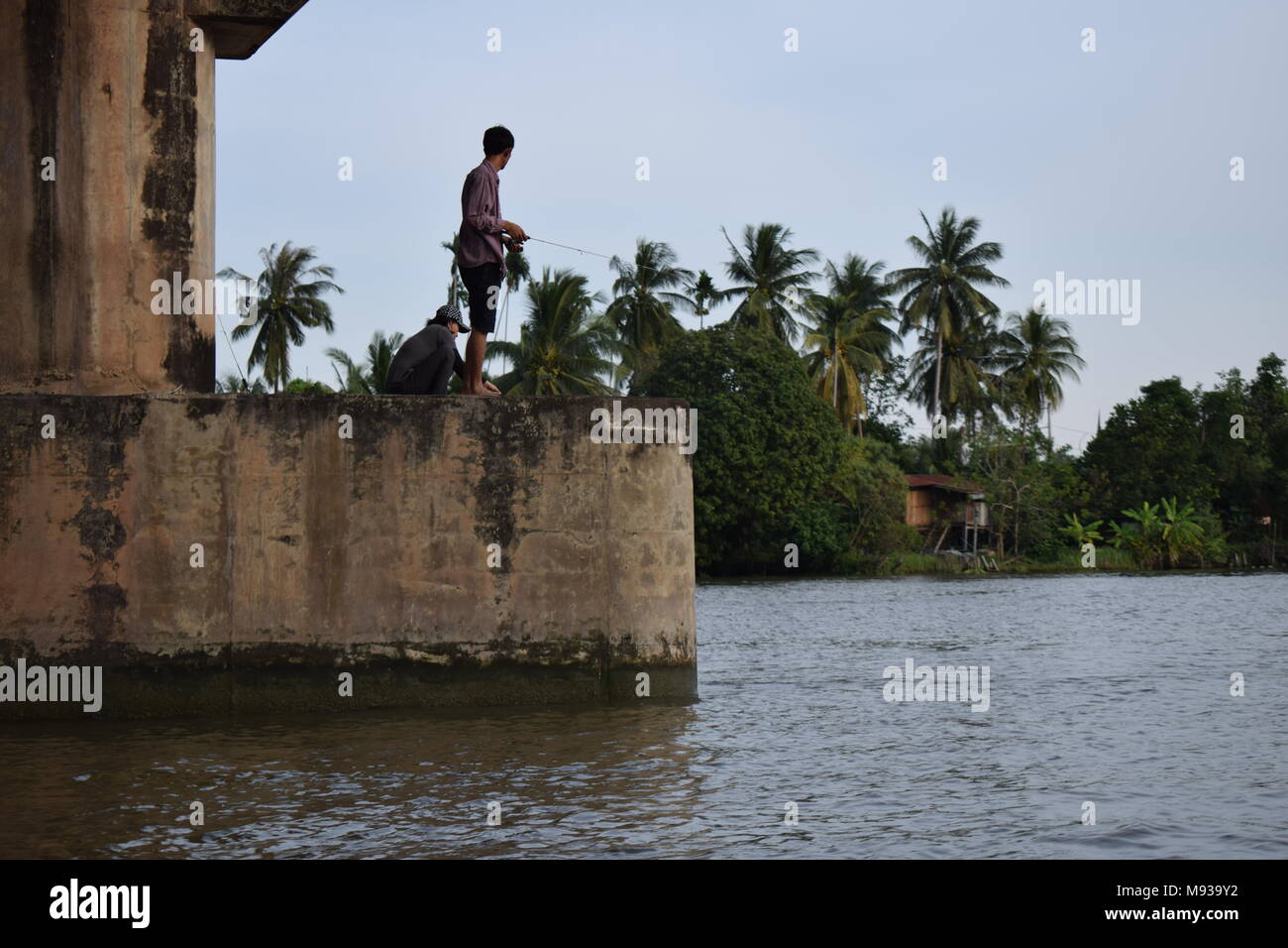 2 La pêche des pêcheurs sous un grand pont sur la rivière Mae klong en Thaïlande prises à partir d'un bateau de tourisme tour avec ciel bleu et de hauts palmiers. Banque D'Images