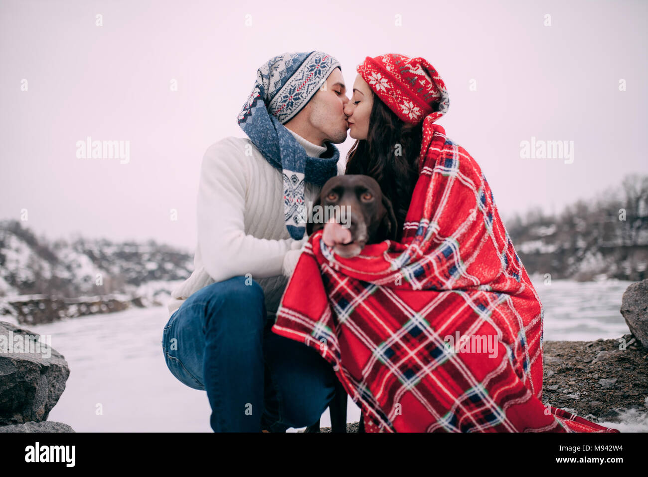 Jeune couple en chapeaux et écharpes avec chien enveloppé dans red plaid  sont assis et s'embrasser à côté de lac gelé et des roches pendant l'hiver  à pied Photo Stock - Alamy