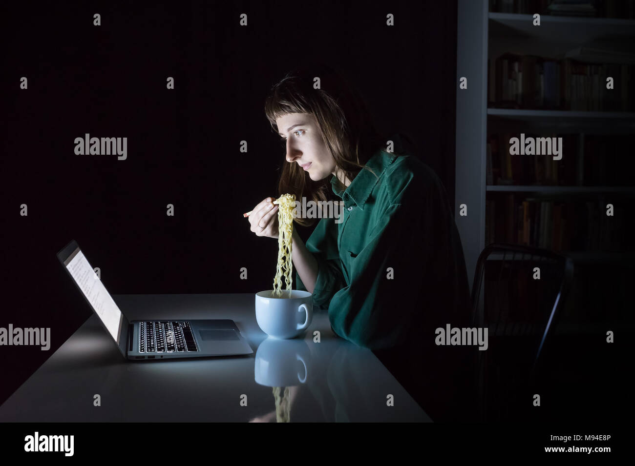 Jeune femme at laptop computer mange nouilles ramen instantané avec des baguettes tard dans la soirée. Femme travaillant ou étudiant en ligne des heures supplémentaires à la nuit a Banque D'Images