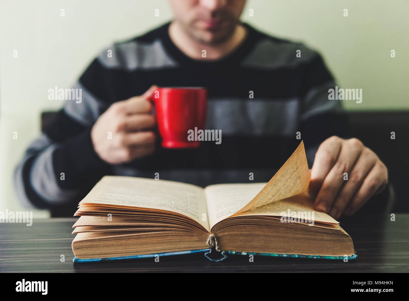 L'homme en pull rayé noir assis par table en bois et de la lecture livre tout en maintenant mug. Routine du soir pour la lecture de livres et d'une tasse de thé dans le même Banque D'Images