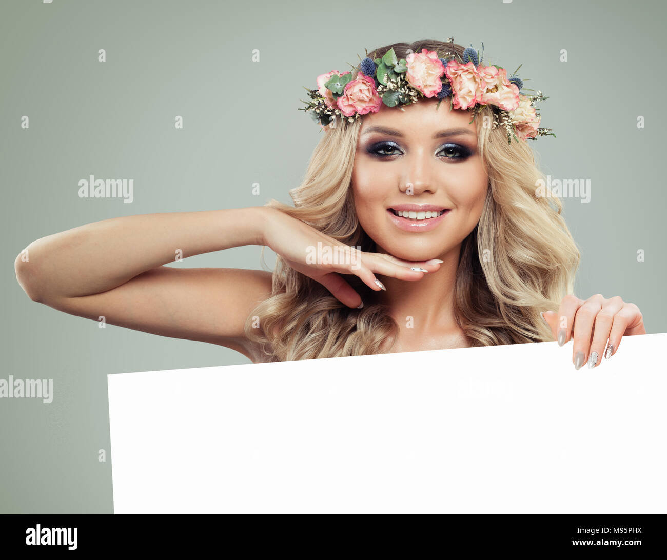Happy Young Woman with flowers Hairstyle Holding Bannière blanche pour le texte. Blonde beauté. Modèle de mode avec de longs cheveux bouclés permanentés et maquillage parfait. S Banque D'Images