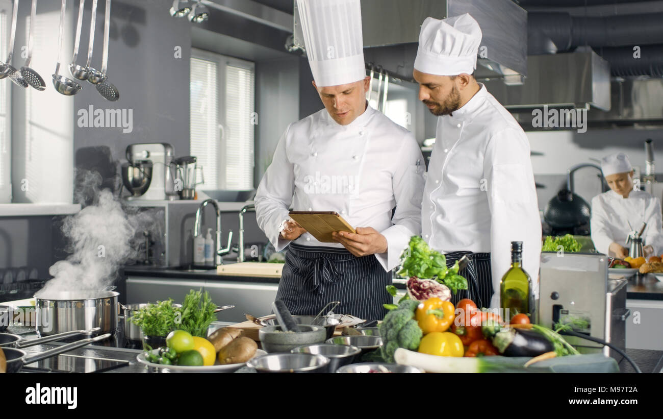 Deux grands Chefs discuter de leur blog vidéo lors de l'utilisation de l'ordinateur tablette. Ils travaillent sur un grand restaurant cuisine professionnelle en acier inoxydable. Banque D'Images