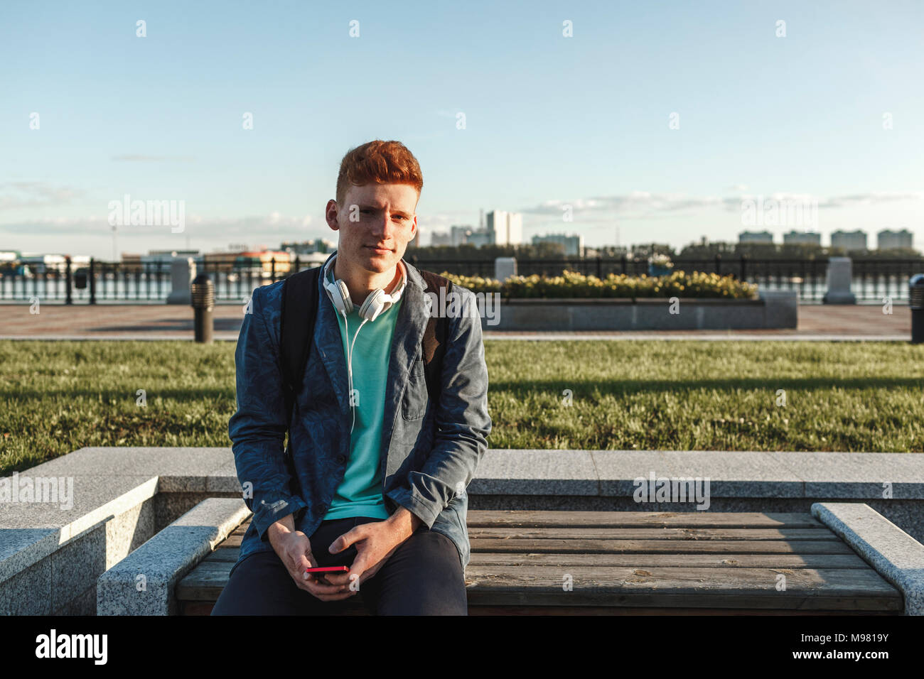 Portrait de jeune homme roux assis sur un banc avec le smartphone et écouteurs Banque D'Images