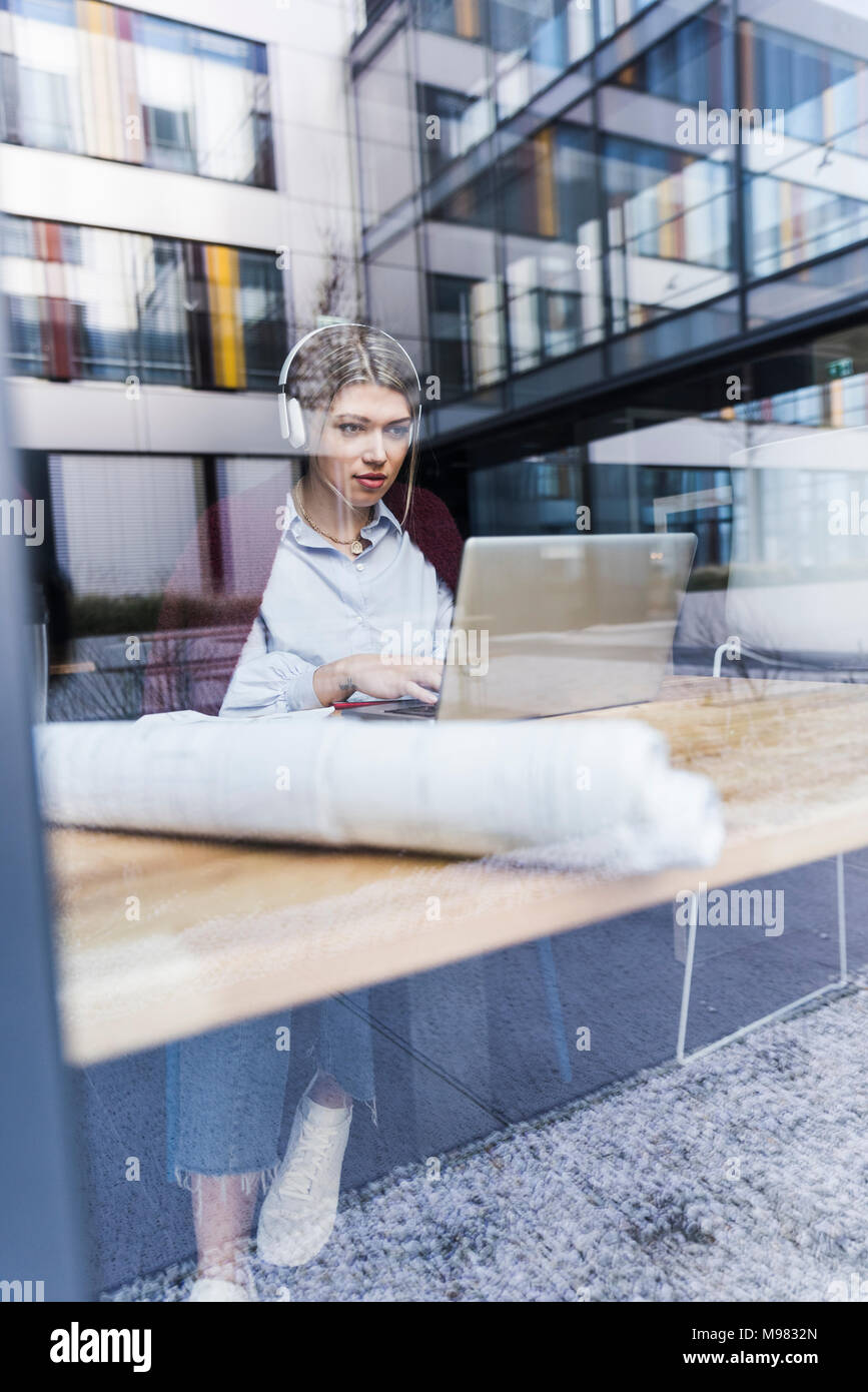 Young woman wearing headphones using laptop derrière la vitre Banque D'Images