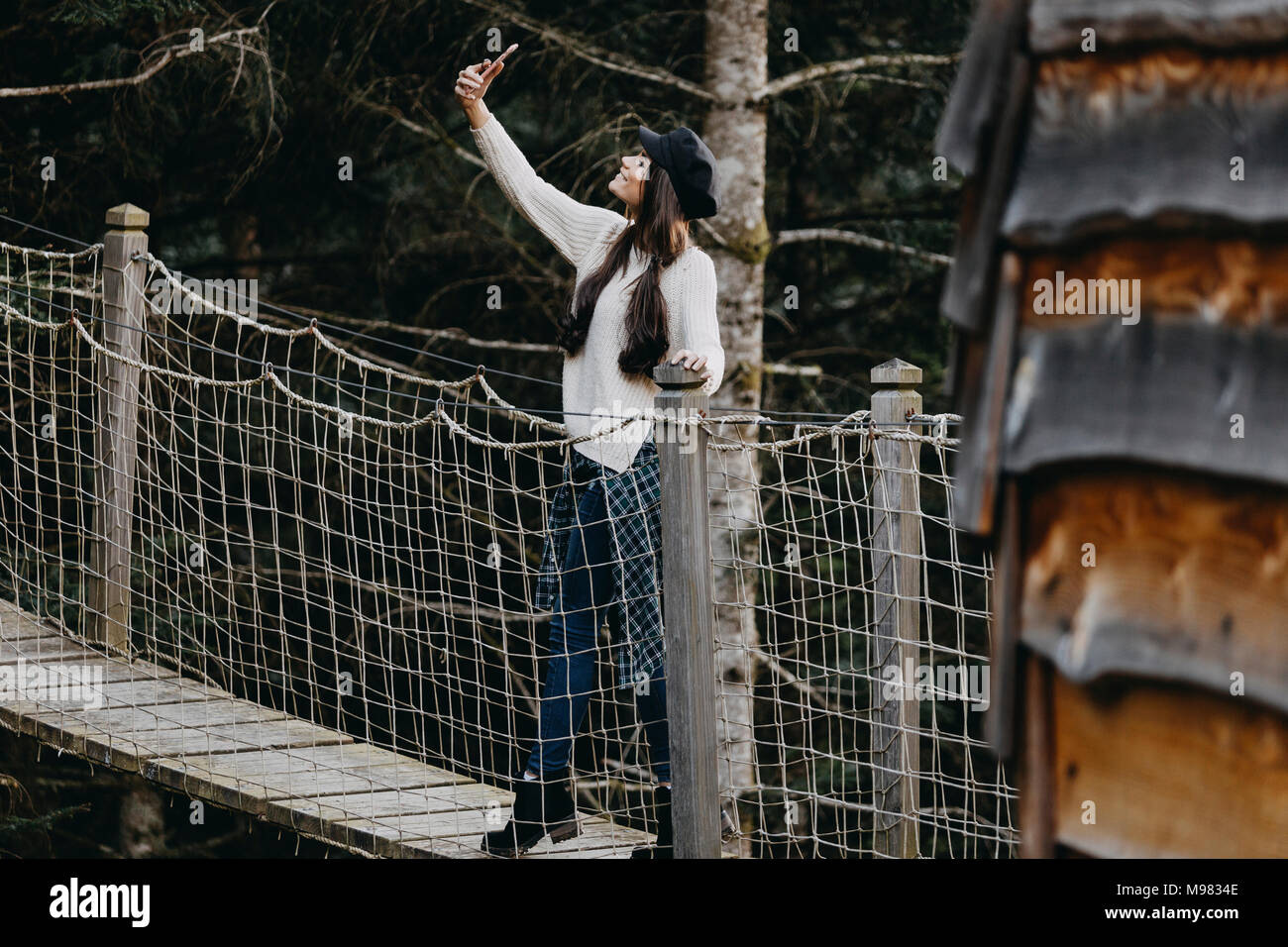 Jeune femme sur un pont suspendu à la maison de l'arbre dans la forêt en prenant un selfies Banque D'Images