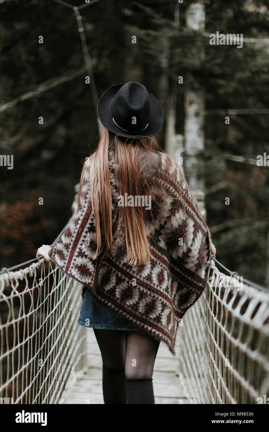 Vue arrière de la mode jeune femme portant chapeau et poncho marche sur suspension bridge Banque D'Images