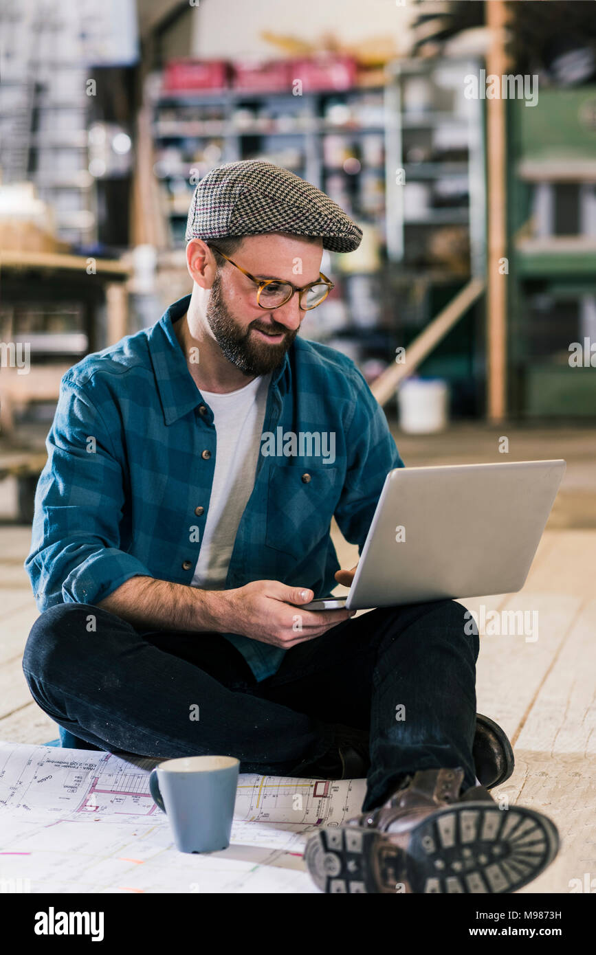 Smiling man with laptop and construction plan assis sur le plancher Banque D'Images