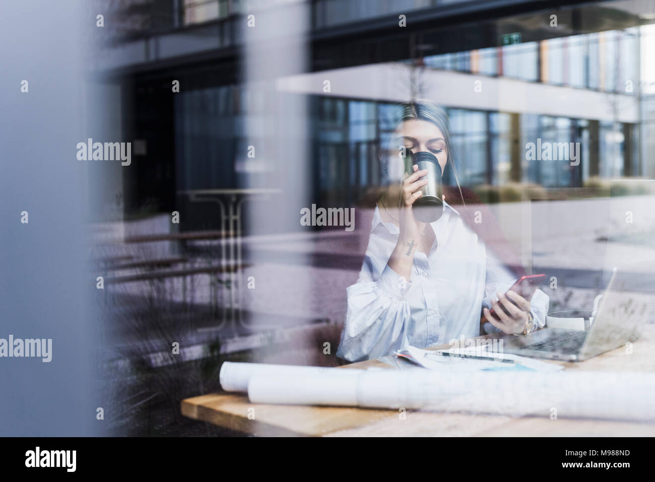 Jeune femme avec un ordinateur portable, téléphone cellulaire et documents de boire du café derrière la vitre Banque D'Images