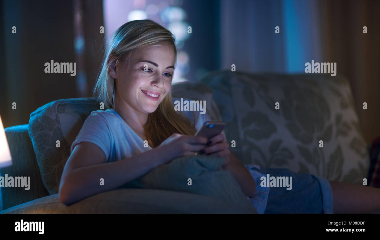 Tard dans la nuit belle femme couchée sur le canapé dans le salon, en utilisant son Smartphone et souriant. Son appartement a grande ville vue de la fenêtre. Banque D'Images