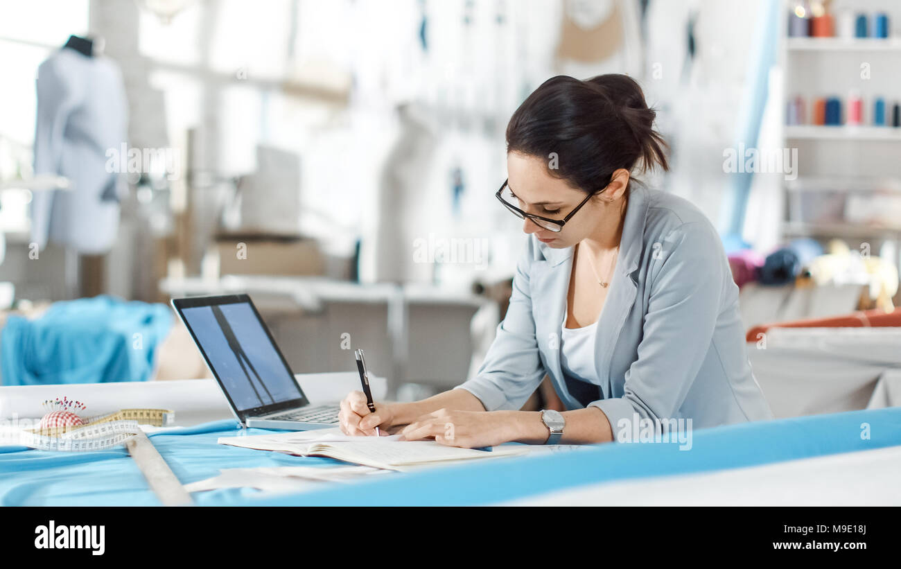 Beau la mode, designer, est assis à son bureau, utilise le smartphone. Son studio est ensoleillée, pleine de détails colorés, tissus, articles de couture, de croquis. Banque D'Images