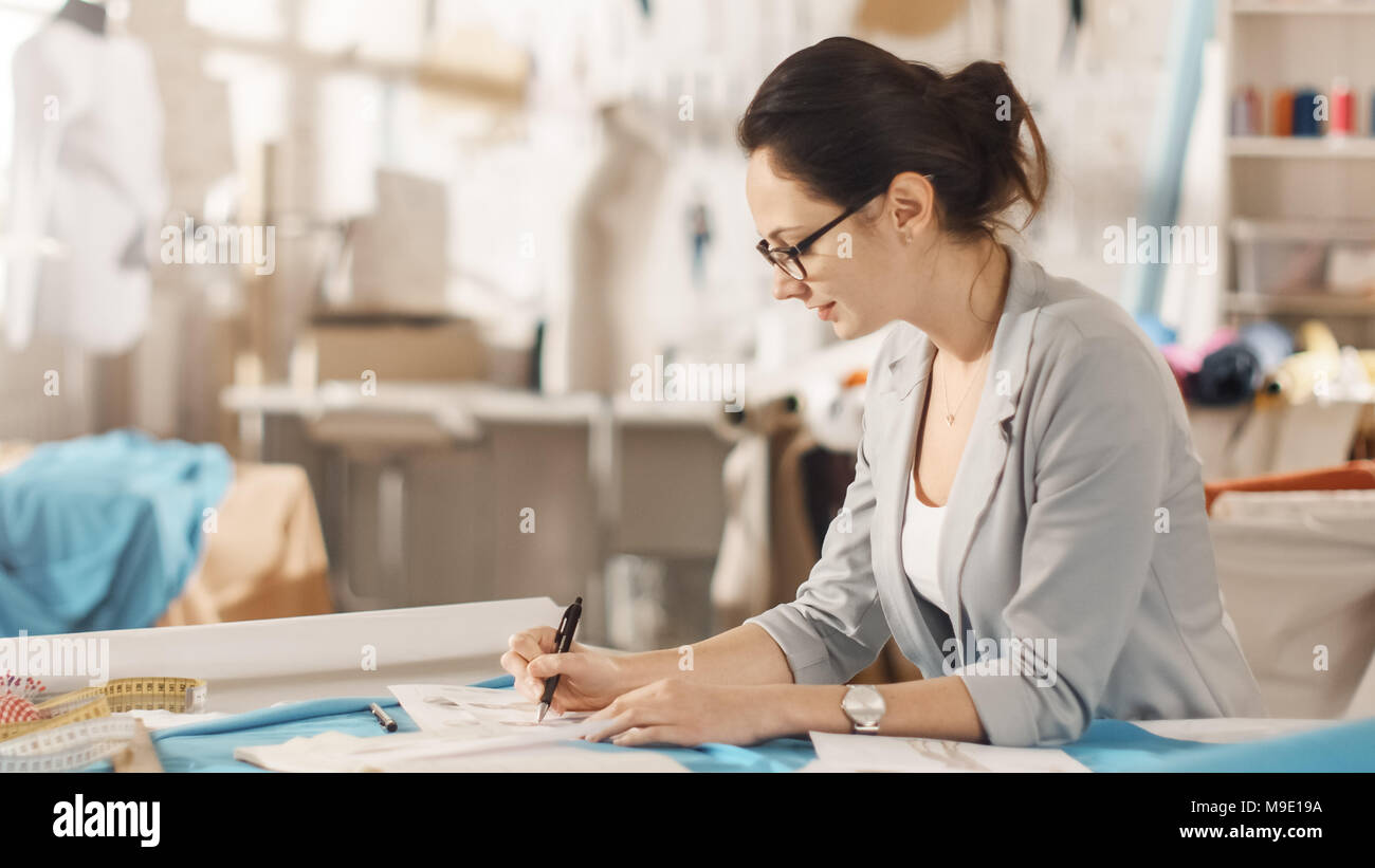 Belle femme la mode, designer, est assis à son bureau et s'inspire des croquis. Son studio est ensoleillée, pleine de détails colorés, tissus, articles de couture, Sketch Banque D'Images