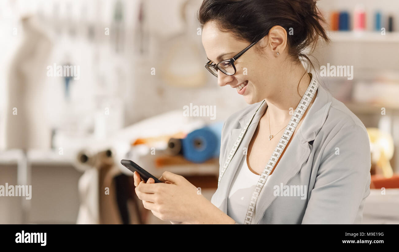 Beau la mode, designer, est assis à son bureau, les types sur le smartphone. Son studio est ensoleillée, pleine de détails colorés, tissus, articles de couture, Sketch Banque D'Images