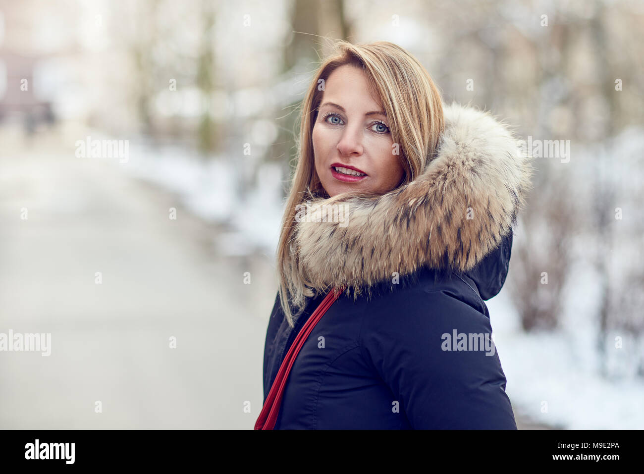 Attractive blonde femme portant une veste chaude avec capuche doublée de  fourrure à l'extérieur permanent en hiver sur une route enneigée en se  tournant vers l'arrière de l'appareil photo Photo Stock -