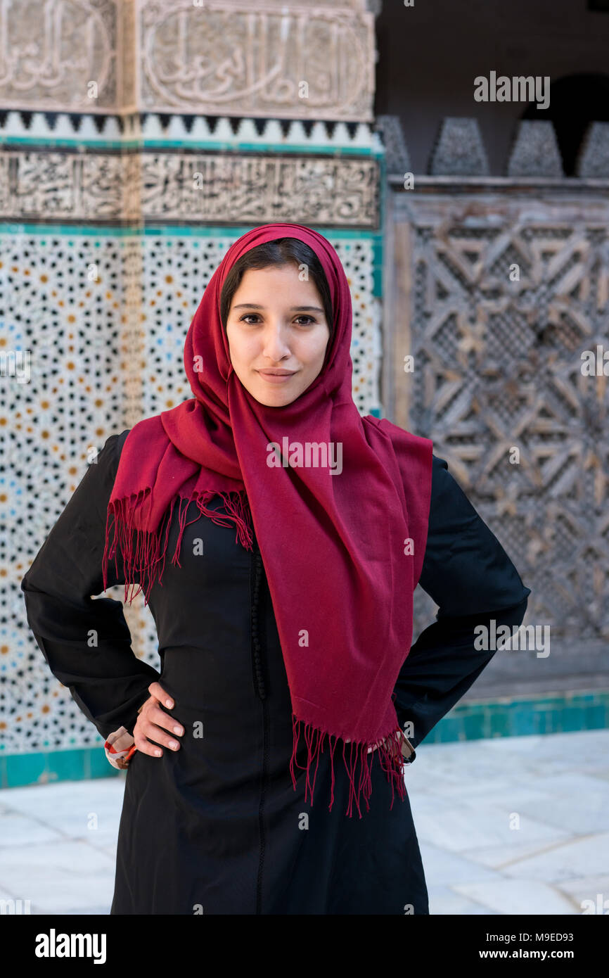 Femme musulmane posant dans des vêtements traditionnels avec hijab rouge et robe noire en face de mur décoré arabesque traditionnels Banque D'Images