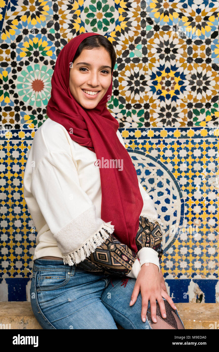 Jeune femme musulmane en jeans avec hijab rouge souriant en face de mur décoré arabesque traditionnels Banque D'Images