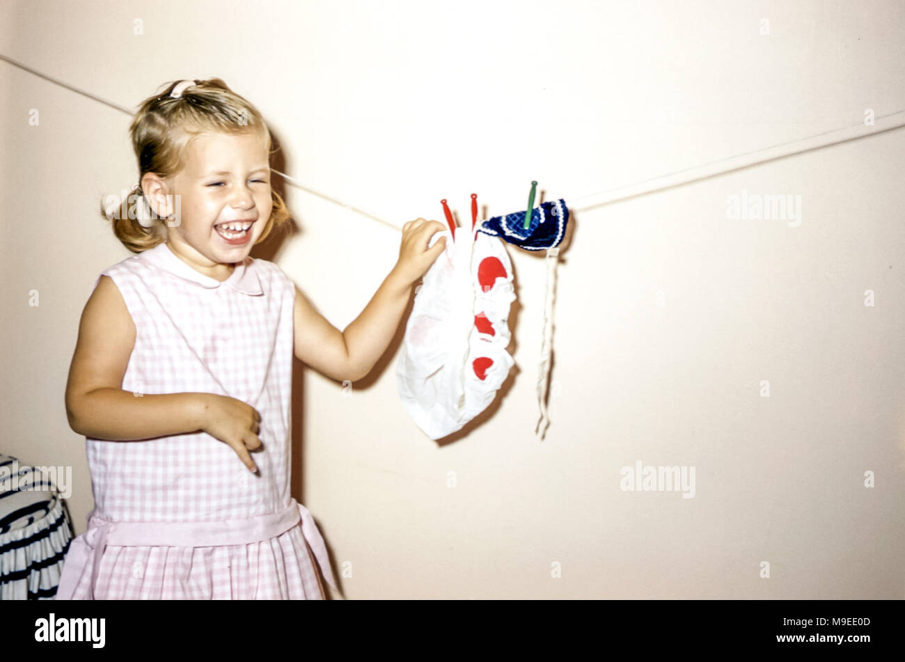 blonde de 2 à 3 ans cheveux jeune fille portant une robe à carreaux roses riant en jouant en famille Maison pendante les vêtements de poupée sur une ligne de lavage aux États-Unis dans les années 1950 Banque D'Images