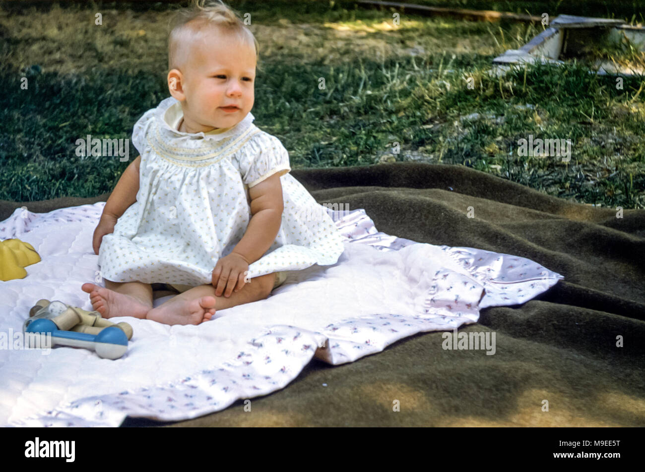 Bébé fille blonde aux cheveux de 4 à 5 mois portant une robe à smocks assise sur un tapis de pique-nique dans un jardin avec des hochets à jouets, San Jose, Californie, États-Unis dans les années 1950 Banque D'Images
