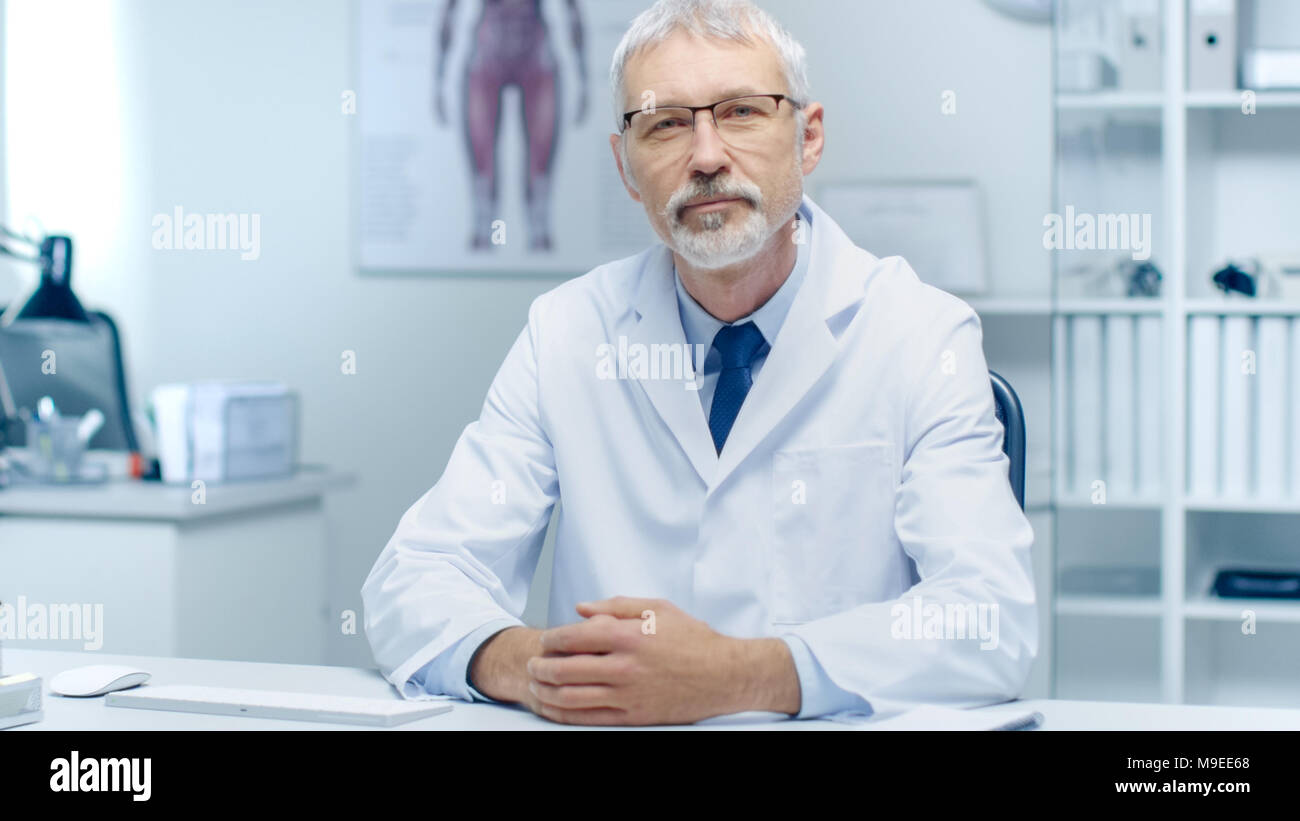 Aux cheveux gris expérimenté Médecin principal et à la caméra en souriant. Portrait en lumière moderne et bureau. Banque D'Images