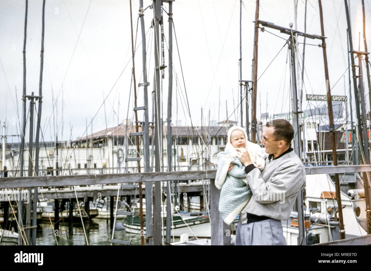 Jeune homme tenant une petite fille de 4 à 5 mois enveloppé dans un châle debout à côté de mâts de bateaux, Fisherman's Wharf, San Francisco Bay, Californie, États-Unis dans les années 1950 Banque D'Images