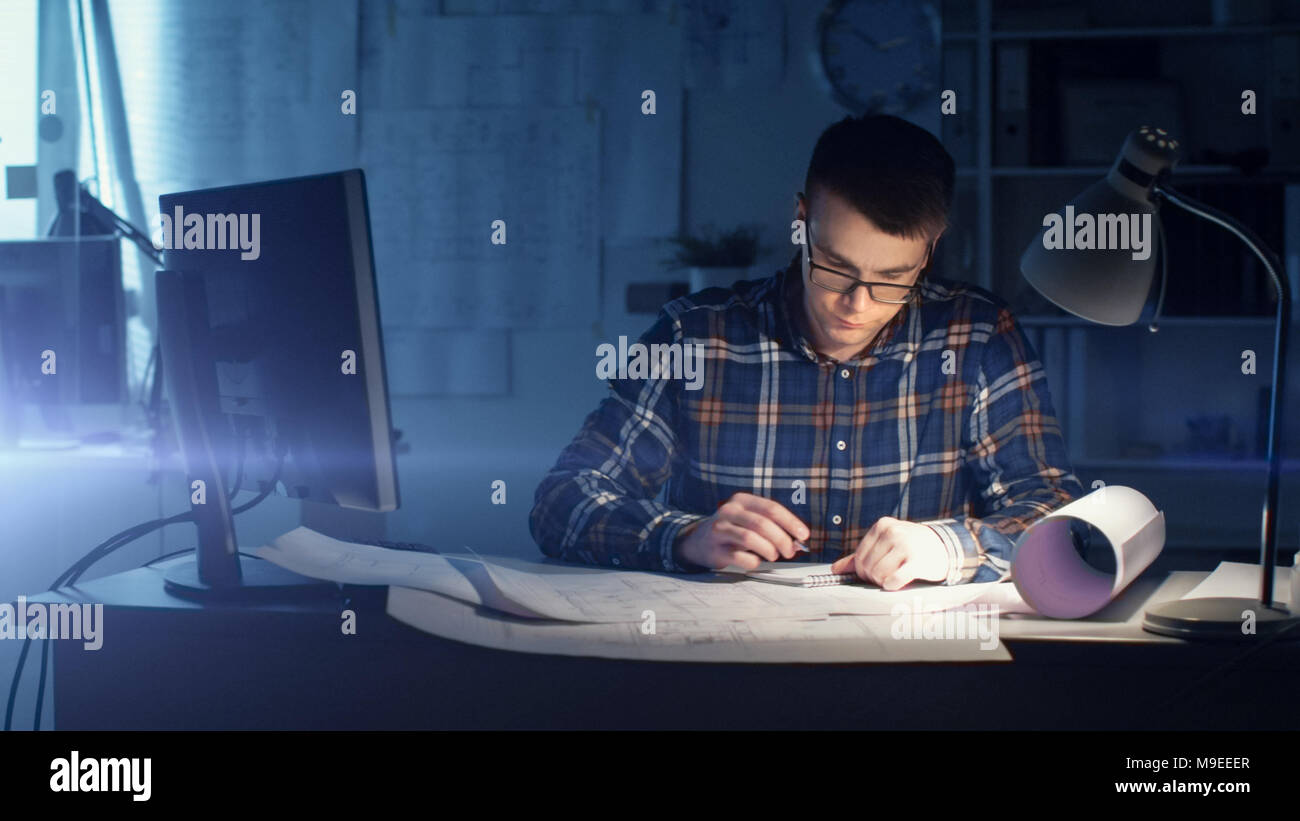 Jeune ingénieur civil architecte capable de travailler de nuit dans son bureau. Office est sombre seulement sa Table s'allume. Divers plans et devis Banque D'Images
