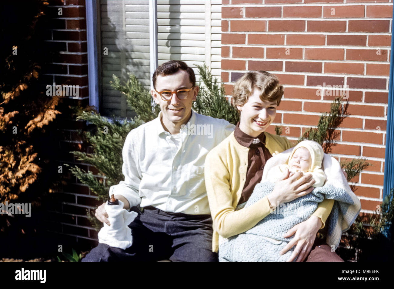 Jeune couple assis au soleil à l'extérieur souriant. La jeune femme tient une petite fille de nouveau-né enveloppée d'un châle et le jeune homme tient une bouteille de bébé, aux États-Unis dans les années 1950 Banque D'Images