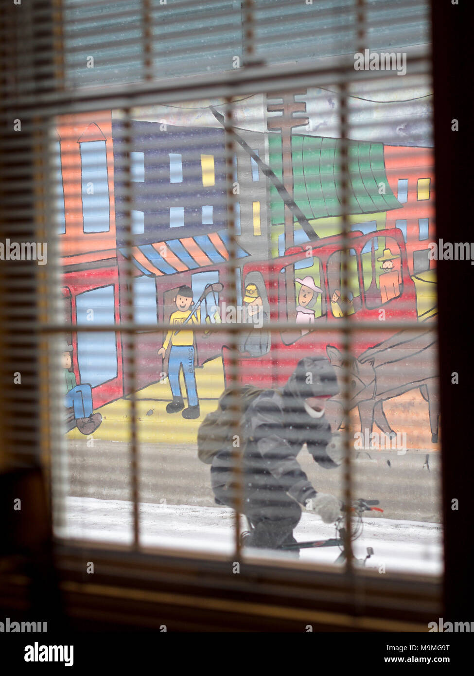 Un cycliste rides par une peinture murale lumineuse comme vu à travers une fenêtre : Une peinture murale de style naïf décore un mur sur une petite rue de la ville de Québec. Un cycliste rides par vus à travers une fenêtre aveugle, peinait sur un jour froid neigeux. Banque D'Images