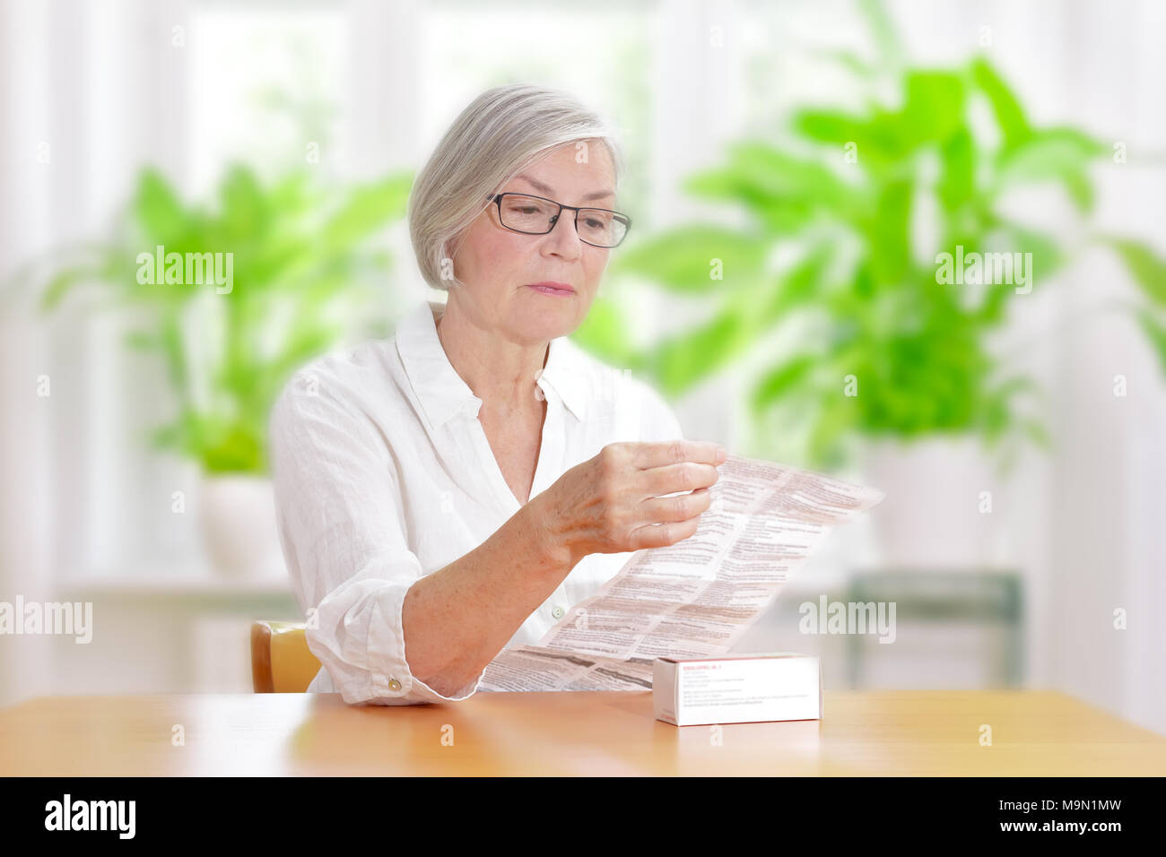 Senior woman sitting at a table dans son salon lire la feuille de renseignements de ses médicaments prescrits Banque D'Images