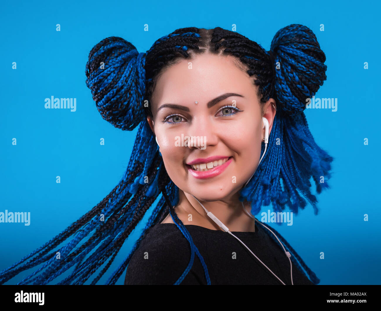 Magnifique jeune femme aux tresses africaines teints hairstyle listening music dans les écouteurs et le chant sur fond bleu. Fille charmante dans écouteurs blancs cheveux dansant avec forme et les yeux fermés. Banque D'Images