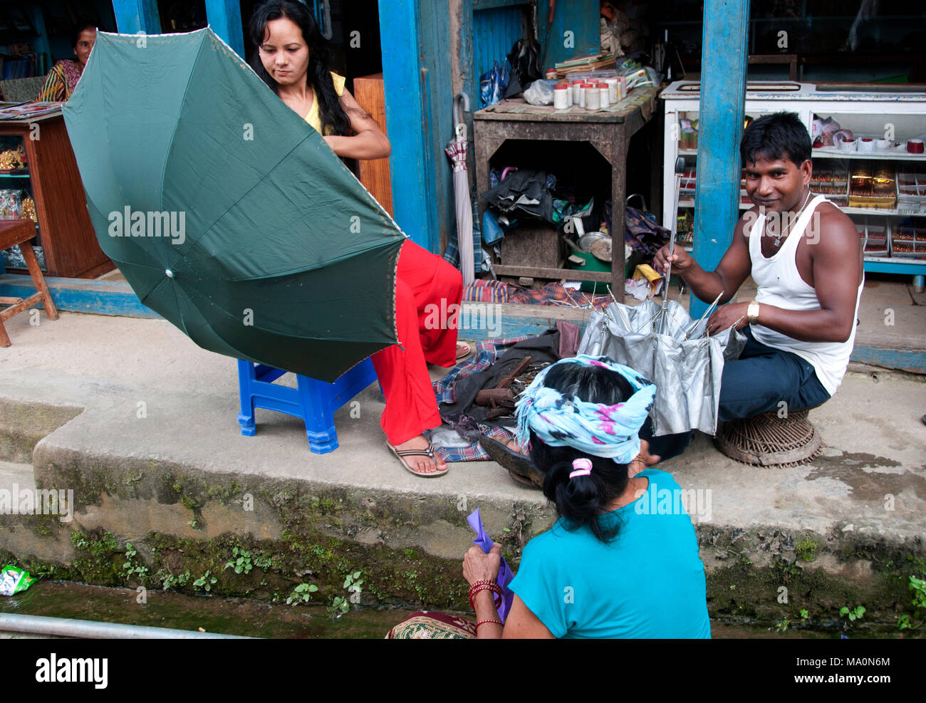 Népal 2014. Khandbari. Umbrella mender travaille sur son terrain de la chaussée en face d'une boutique, avec des clients en attente. Banque D'Images