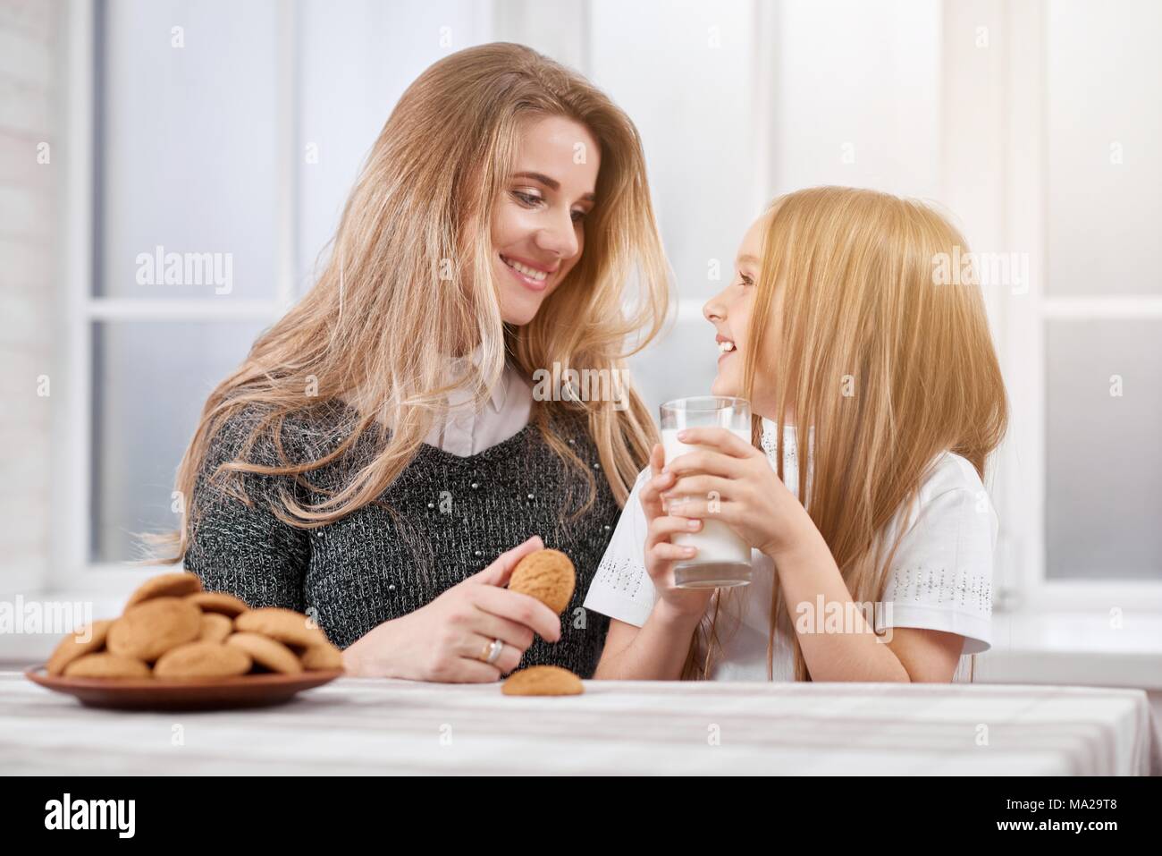 Photo de deux sœurs plus jeunes et rire-Oder. Les deux filles sont blonds cheveux raides. Ils mangent de délicieux cookies de plaque et boire du lait. Les soeurs s'asseoir sur le livre blanc arrière-plan de la fenêtre. Banque D'Images