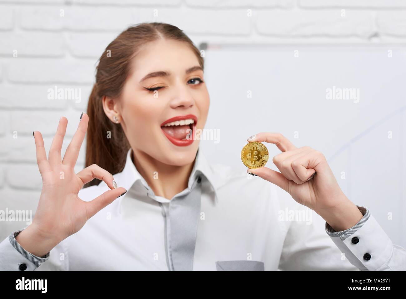 Attractive young model holding golden comme bitcoin cryptocurrency principal et montrant ok s'identifier tout en clignant de l'espièglerie à l'appareil photo. Le succès de capture horizontale professionnelle féminine, arrière-plan flou. Banque D'Images