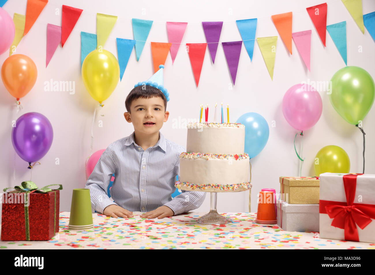 Petit garçon avec un chapeau de fête et un gâteau d'anniversaire contre un mur avec des drapeaux et des ballons de décoration Banque D'Images