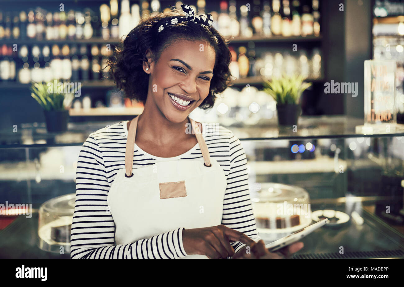 Souriante jeune entrepreneur africain portant un tablier et à l'aide d'une tablette numérique alors qu'il se tenait devant le comptoir d'un café à la mode Banque D'Images