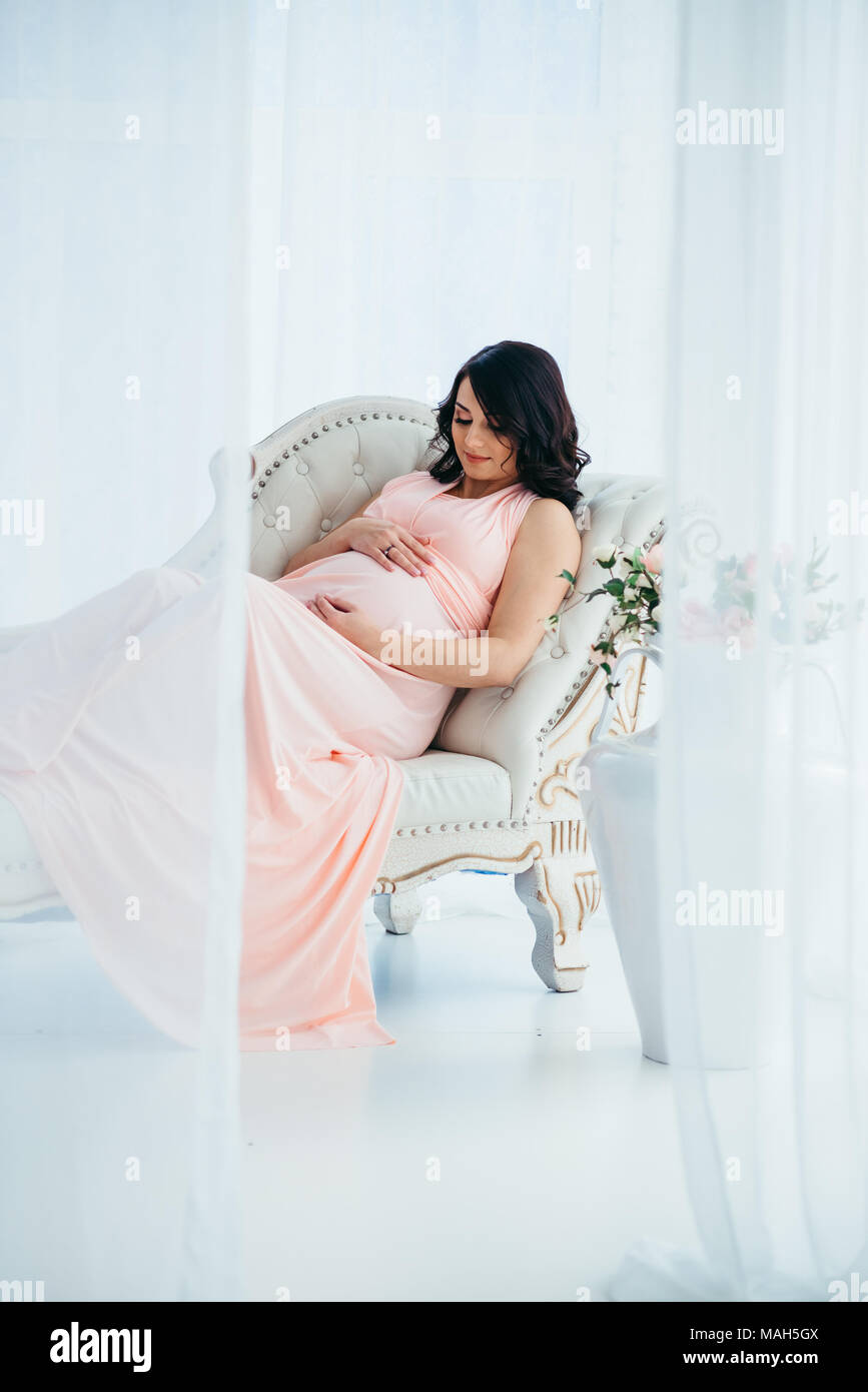 La jolie charmante femme enceinte en robe rose longue est de toucher son ventre en position allongée sur le canapé près de vase avec fleurs dans moder studio. Banque D'Images