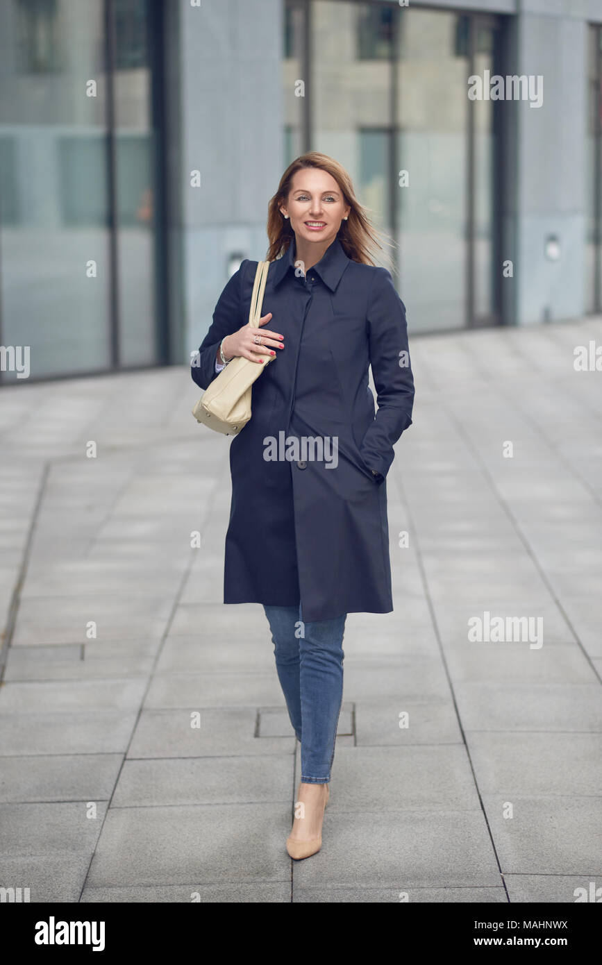 Belle blonde élancée élégante femme en jeans et d'un pardessus de marcher le long d'une rue urbaine à la voiture sur le côté avec un sourire Banque D'Images