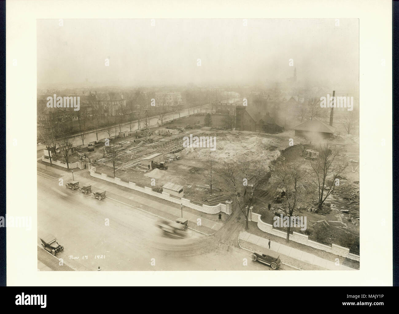 Photographie sépia, horizontales de chantier sur un coin de rue. Le site est entouré par une clôture en pierre blanche, et les travaux de fondation sont en cours. La stabilité de la résidence William K. Bixby est encore visible à l'arrière du site. Titre : Vue aérienne de la construction à l'emplacement de l'hôtel Chase, 17 novembre 1921. . 17 novembre 1921. Banque D'Images