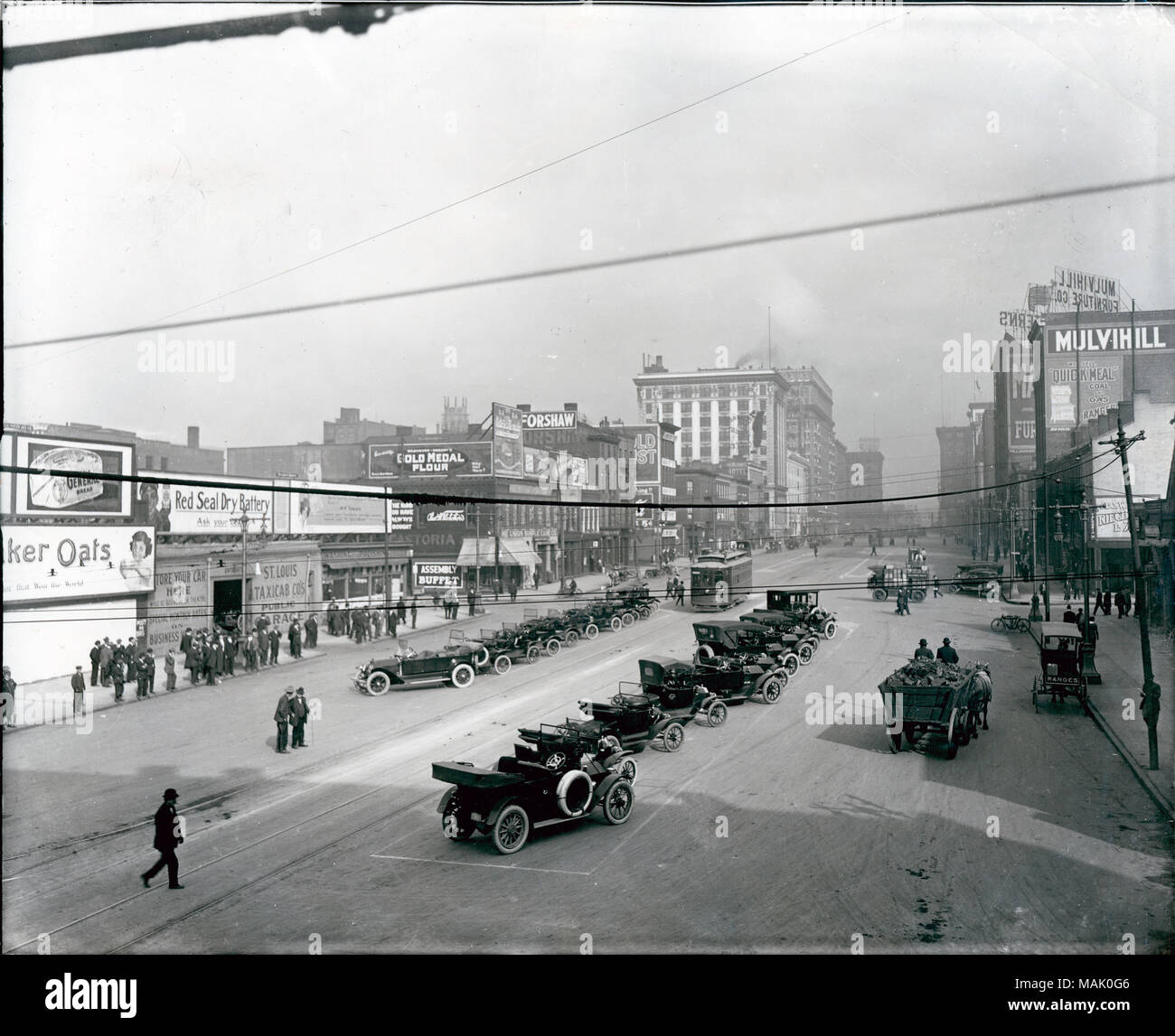 À l'horizontal, photographie en noir et blanc d'une vue sur la rue de la 12e Rue. Un voyage à travers les rangées de voitures en stationnement. Ligne de personnes le trottoir le long d'une rangée de bâtiments qui comporte des annonces publicitaires pour "Quaker Oats", "pile sèche du Sceau rouge', 'St. Louis Co Taxi's Garage public', 'Assemblée générale Buffet', 'OR', 'Farine Forshaw', 'l'Union européenne Exemple Co.',« mulvihill" et "repas rapide'. Titre : Centre de la rue parking automobile le tramway lignes pistes sur Douzième Street, à au nord de châtaignier. . Vers 1915. Swekosky, William G., 1895-1964 Banque D'Images