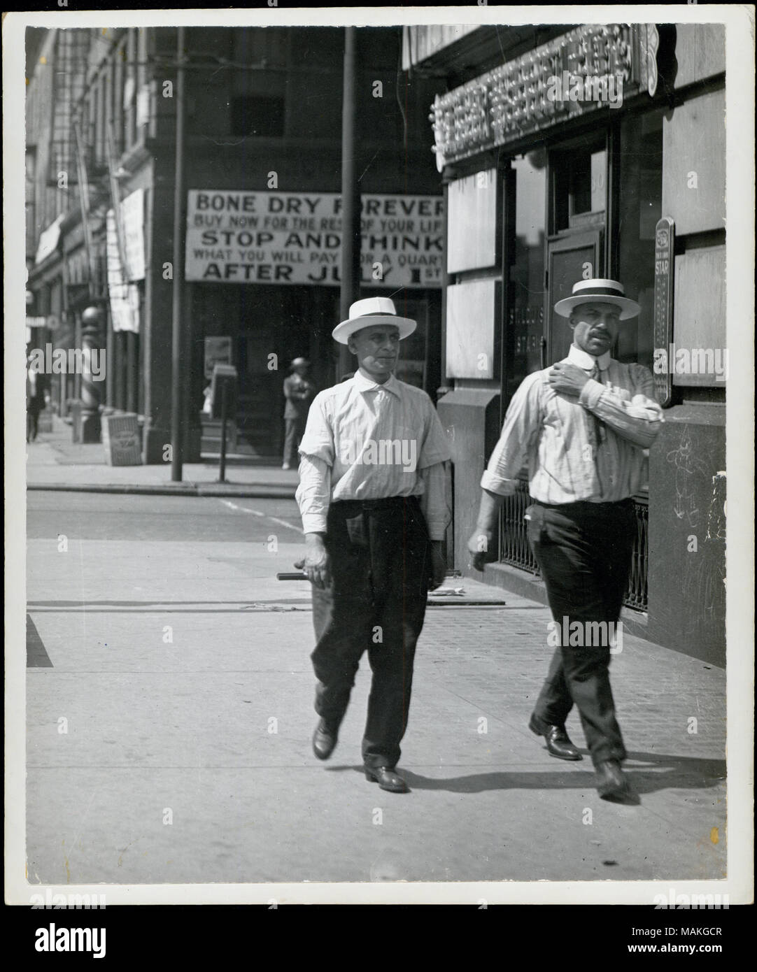 Vertical, photographie noir et blanc montrant deux hommes chemise en marchant sur le trottoir. Derrière eux, un grand signe taverne proclame l'imposition de la première phase de l'interdiction. Il y est question de "archi-sèche pour toujours / achetez pour le reste de votre vie / arrêter et penser ce que vous allez payer pour un quart après le 1er juillet.' Title : deux hommes en chemise en juin 1919. Tavern signe en angle sud-ouest de douzième et Olive proclame l'imposition imminente de la première phase de l'interdiction. . Juin 1919. Banque D'Images