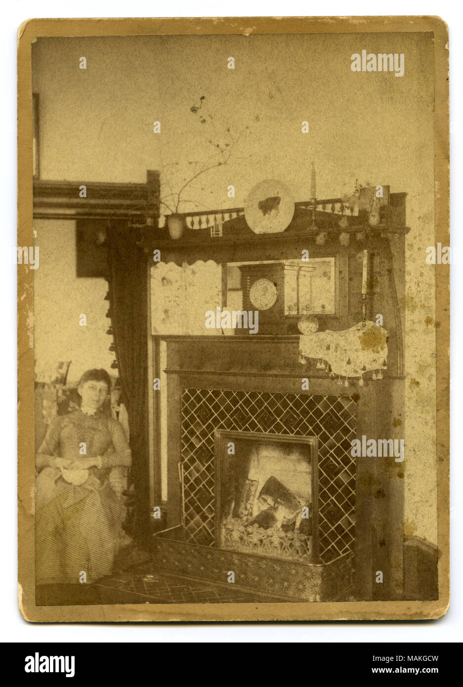 Photographie sépia, verticale montrant une femme assise à la gauche d'une cheminée. Il y a de nombreux articles sur le manteau dont un réveil, de la direction générale dans un vase, une assiette, et deux chandeliers. Titre : Femme assise à côté d'une cheminée allumée. . Vers 1880. Banque D'Images
