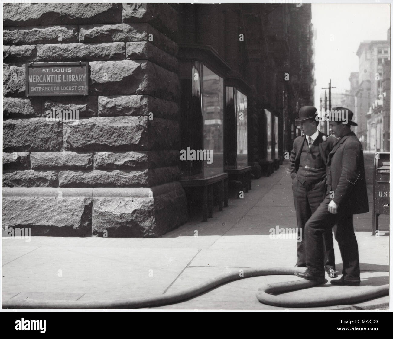 L'horizontale, 1970 Photographie noir et blanc montrant le coin de la Saint-louis bibliothèque. Mercantile Un signe sur l'édifice se lit comme suit : 'St. Bibliothèque Louis Mercantile / Entrée sur Locust St.' plusieurs cas d'affichage peut être vu le long du côté du bâtiment, s'étendant dans l'arrière-plan. Deux hommes debout sur le coin de rue pipes. L'un des hommes repose son pied sur un grand flexible qui serpente le long du trottoir au bord inférieur de l'image. Titre : coin de la Saint Louis, bâtiment Bibliothèque Mercantile 510 Locust Street. . Entre 1900 et 1920. Holt, Charles Clement, 1866-1925 Banque D'Images