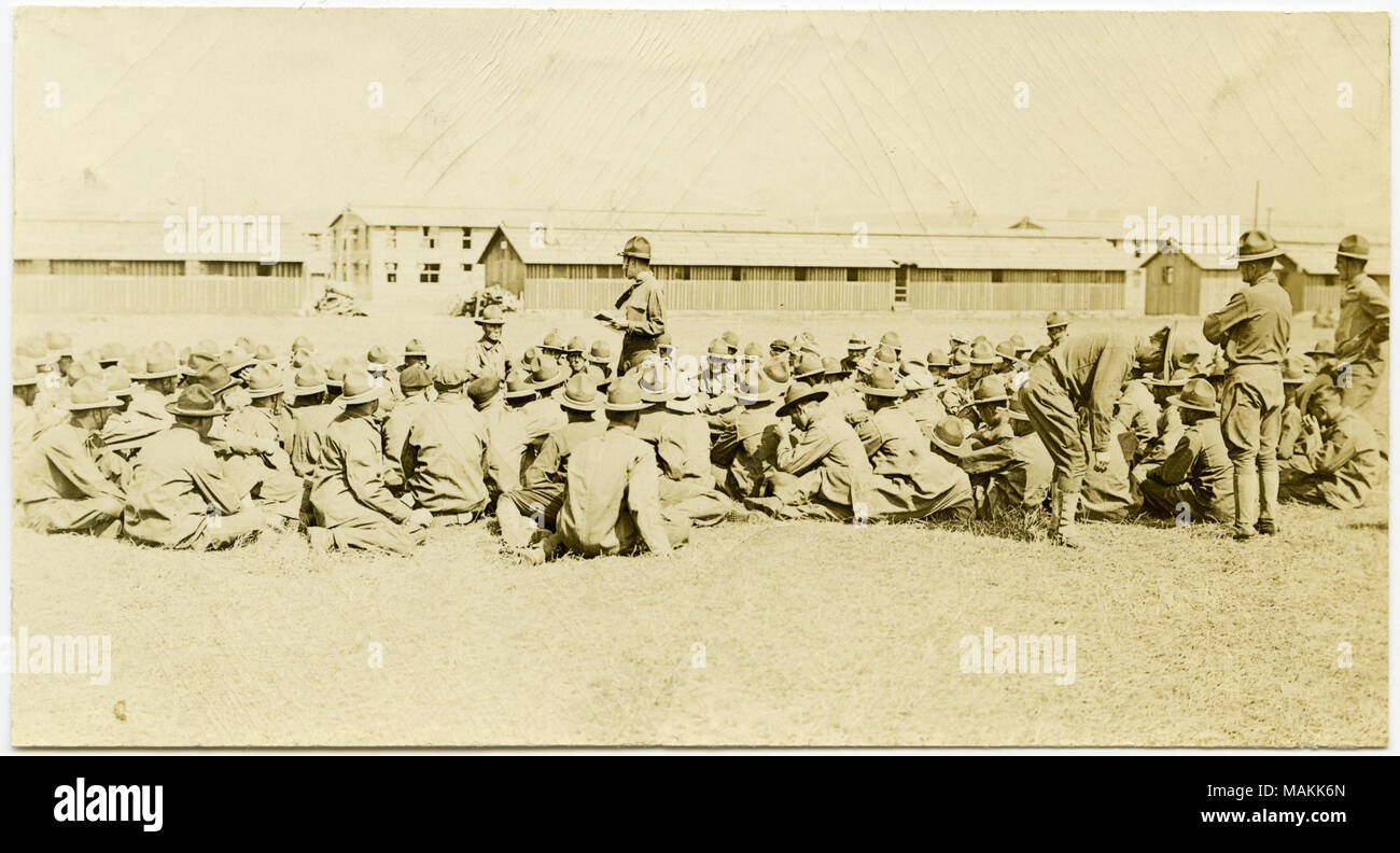 L'horizontale, sépia photographie montrant un grand groupe de soldats en uniforme assis sur le sol dans un champ d'herbe tandis qu'un soldat se trouve dans le centre du groupe de parler. Divers bâtiments peut être vu dans le contexte de l'image. Titre : Les soldats assis sur le sol dans un groupe à l'écoute de l'un soldat dans le centre de parler. . Entre vers 1914 et vers 1918. Michel, Carl Banque D'Images