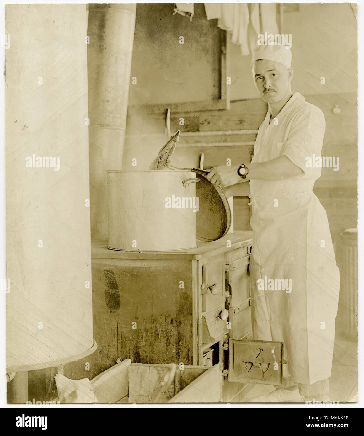 Photographie sépia, verticale montrant un homme en civil portant un tablier, debout devant une cuisinière dans une cuisine. L'homme est à l'aide d'une fourche à deux volets pour extraire un élément d'une grande casserole assis sur la plaque de cuisson. Titre : Man working in Kitchen tire un élément dans un grand pot sur une plaque de cuisson. . Entre vers 1914 et vers 1918. Michel, Carl Banque D'Images