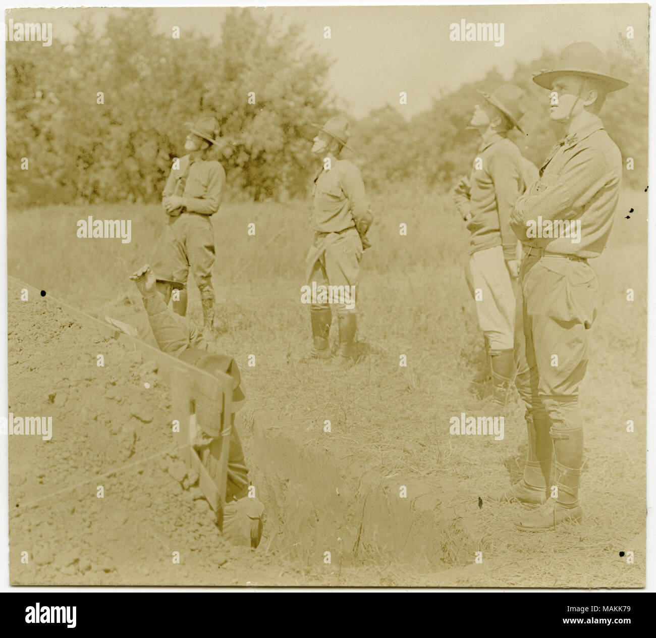 Photographie sépia, Horizontal montrant des soldats en uniforme qui participent à un exercice d'entraînement. Cinq soldats sont sur le terrain à la recherche vers quelque chose dans le ciel. En face d'eux a au moins deux soldats debout dans une tranchée peu profonde. Les hommes sont debout dans un champ d'herbe, et des arbres peut être vu dans la distance. Titre : des soldats dans une tranchée peu profonde qui participent à un exercice de formation alors que d'autres observent. . Entre vers 1914 et vers 1918. Michel, Carl Banque D'Images