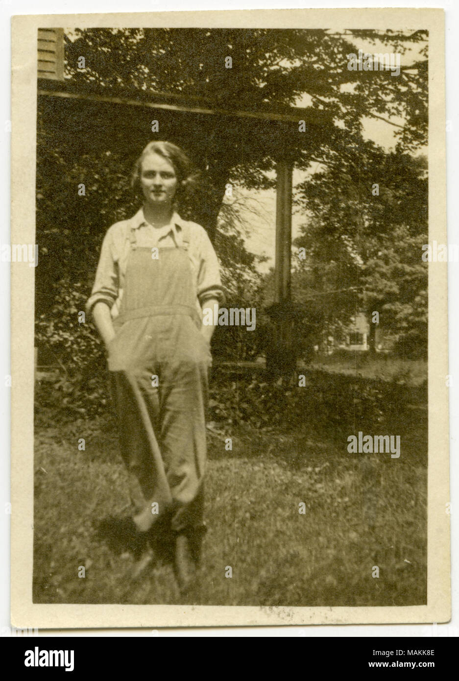 Noir et blanc, photo à orientation verticale d'une jeune femme portant des bretelles dans une ferme. La femme est un membre de l'Ohio Femme's Club. De nombreux arbres apparaissent tout autour en arrière-plan, et porche-comme-faux d'une maison apparaît immédiatement derrière elle. Une autre chambre est à l'arrière-plan à droite très lointain. Retour de la photo se lit, manuscrit, "l'un des MO. groupe / un '1918' farmerette.' Title : Texas Woman's Club états portant des salopettes. . 1918. Banque D'Images