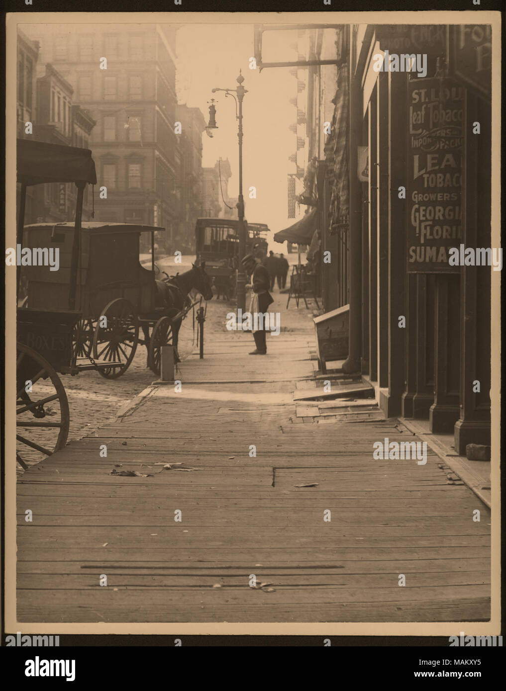 Vertical, photographie en noir et blanc d'un véhicule stationné sur un trottoir la planche en bois. Store fronts line le trottoir et un homme se tient sur le coin. Titre : jusqu'à la rue du marché en bois de trottoir en face de Saint Louis Les importateurs de tabac en feuilles. . Entre vers 1800 et vers 1900. Swekosky, William G., 1895-1964 Banque D'Images