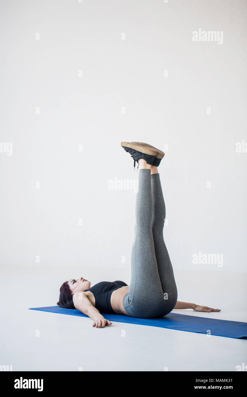 Young attractive female trainer est en train de faire les jambes. La femme athlétique est allongé sur le tapis de yoga et d'élever ses jambes. Location studio. Banque D'Images