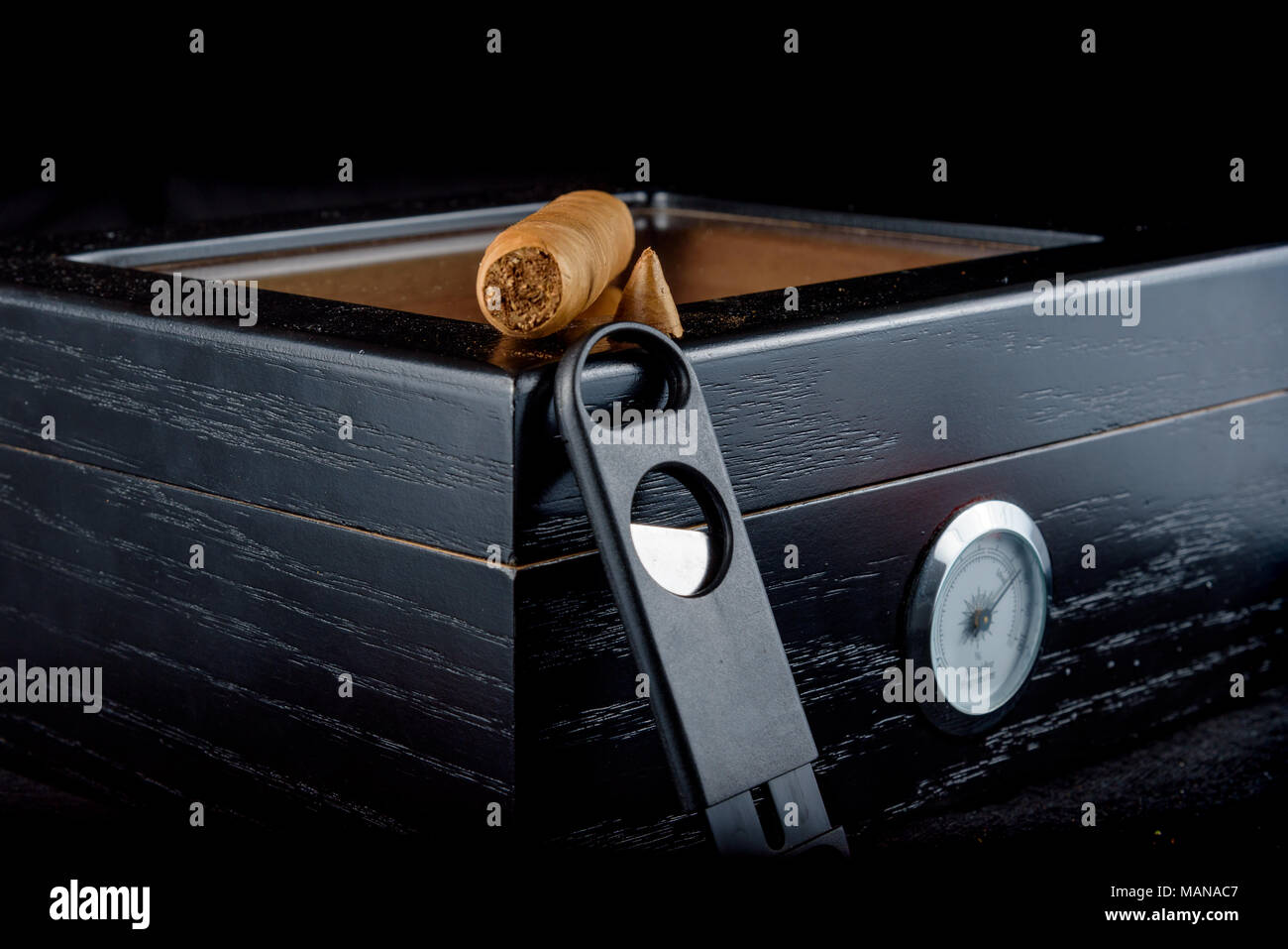 Une riche odeur de cigare qui a été coupe droite avec fumée. Reposant sur un humidor noire à côté d'un coupe-cigare tout droit. Fond noir Banque D'Images
