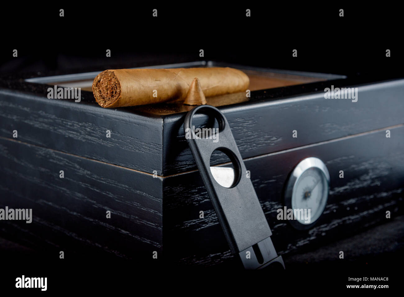 Une riche odeur de cigare qui a été coupe droite avec fumée. Reposant sur un humidor noire à côté d'un coupe-cigare tout droit. Fond noir Banque D'Images