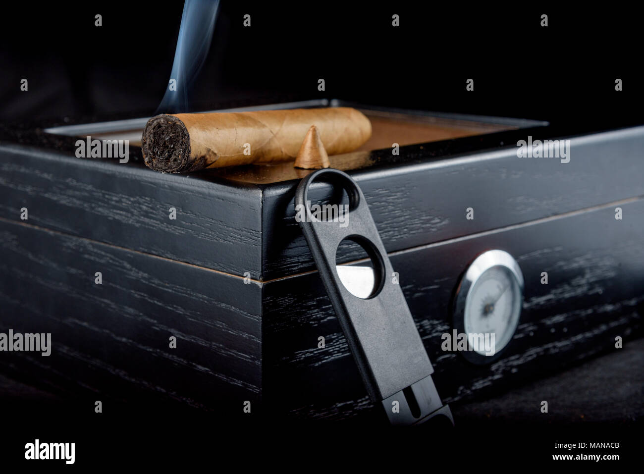 Une riche odeur de cigare qui a été coupe droite avec fumée. Reposant sur un humidor noire à côté d'un coupe-cigare tout droit. Fond noir Banque D'Images