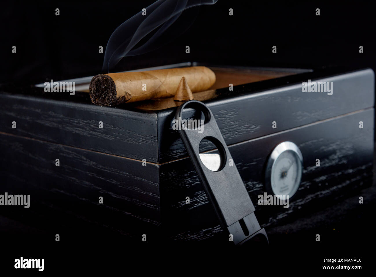 Une riche odeur de cigare qui a été coupe droite avec fumée. Reposant sur un humidor noire à côté d'un coupe-cigare tout droit. Fond noir Banque D'Images